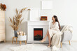 © Pixel-Shot - Young woman on armchair looking at paintings above fireplace in living room