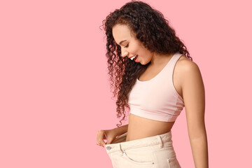 Poster - Surprised young African-American woman in loose jeans on pink background. Weight loss concept