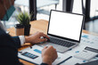 © amnaj - Business professional working on a laptop with a blank screen at a desk, analyzing financial documents and graphs, with a coffee cup and plant.