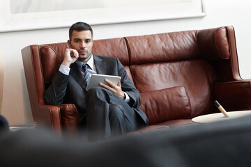 Canvas Print - Businessman, serious and tablet on sofa in office or waiting room for appointment with lawyer in portrait. Worker, digital or professional on couch with tech for consult with browsing online in Spain