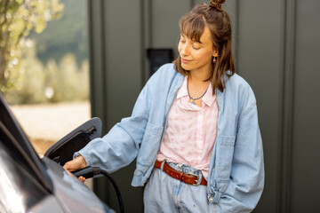 Young woman plugging a charger into electric vehicle near her house outdoors. Concept of modern technologies, EV cars and sustainable lifestyle