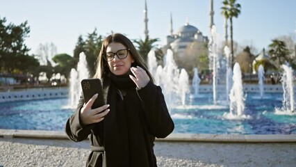 Wall Mural - A smiling woman takes a selfie with her smartphone at sultanahmet park, with the blue mosque and a fountain in istanbul, turkey.