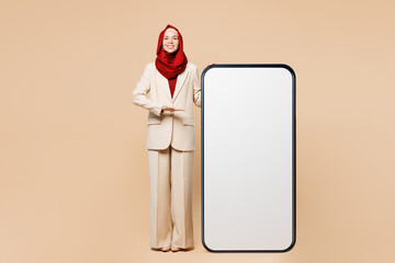 Wall Mural - Young smiling Arabian Asian Muslim woman wear red abaya hijab suit clothes point hand on big huge blank screen area mobile cell phone smartphone isolated on plain beige background. UAE Islam concept.