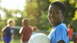 © Felippe Lopes - Young boy holding a soccer ball smiles back at the camera on a sunny day, with other children playing in the background.