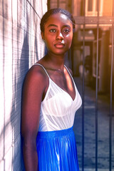 Wall Mural - Portrait of a beautiful Afro-American woman in a white top and blue skirt, standing confidently against a textured wall, urban life concept