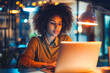 © VisualProduction - A confident young Afro-American woman typing on her laptop in a modern office flooded with natural light, her determined expression illuminated by the glow of her computer screen.