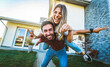 © Davide Angelini - Happy young couple holding home keys after buying real estate - Husband and wife standing outside in front of their new house