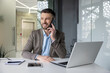 © Liubomir - Smiling businessman talking on the phone while sitting at a desk in a modern office workspace with laptop and notepad.