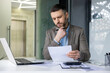 © Liubomir - A businessman deeply focused on analyzing documents at his desk in a modern office. He appears thoughtful and contemplative while holding a pen.