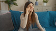 © Krakenimages.com - Beautiful young hispanic woman talking on the phone while sitting on a blue-covered couch in a cozy living room with indoor plants and decorative pillows.