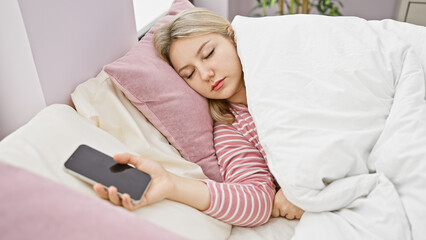 Sticker - A young blonde woman sleeping in a cozy bedroom, holding a smartphone in her hand, depicting a peaceful indoor scene.