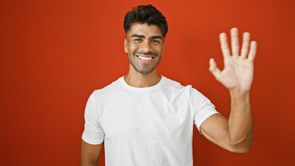 Wall Mural - Handsome young hispanic man with beard waving hello against an isolated red background.