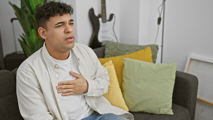 Wall Mural - A young man looks uncomfortable while sitting on a couch in a modern living room with a guitar in the background.