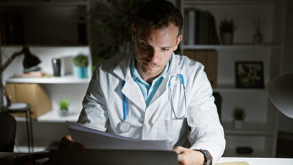 Canvas Print - A focused young hispanic man with a beard wearing a lab coat and stethoscope reviews documents in a clinic office at night.