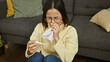 © Krakenimages.com - A young woman in a yellow sweater uses a tissue while holding a thermometer, sitting on a grey sofa indoors.