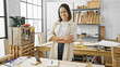 © Krakenimages.com - Hispanic woman smiling confidently in a carpentry workshop surrounded by tools and woodwork.