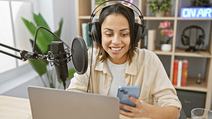 Smiling young woman with headphones in a radio studio holding a smartphone while recording a podcast.