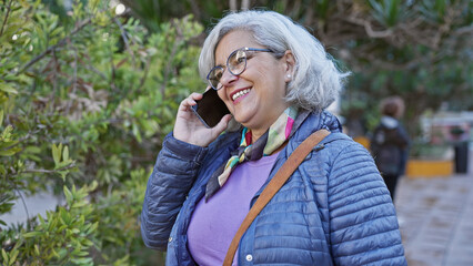 Poster - Cheerful mature woman with grey hair talking on smartphone in a city park, surrounded by greenery.