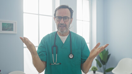 Canvas Print - Mature hispanic man in medical scrubs gesturing uncertainty in a bright clinic room