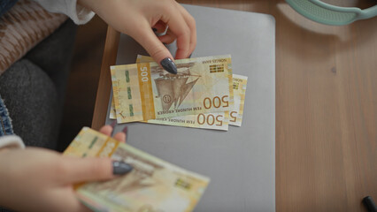 A young caucasian woman counts norwegian kroner at a table indoors, depicting finance and domestic life.