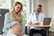 © Home-stock - Portrait of happy expectant lady embracing belly and smiling, sitting at private clinic during consultation with male gynecologist