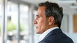 © iuricazac - A man in a suit looking out a window with a thoughtful expression standing in a modern office with a blurred view of greenery and buildings outside.