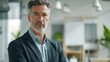 © iuricazac - A man with a beard and gray hair wearing a dark suit and a light blue shirt standing in a modern office with a thoughtful expression.