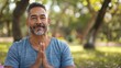 © iuricazac - A man with a beard and mustache wearing a blue shirt sitting in a park with his hands clasped together in a meditative pose.
