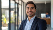 © Maksym Tereshchenko - Smiling elegant confident young professional Latino Hispanic businessman with a polished look, radiating leadership, standing in a bright, contemporary office