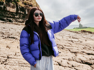 Wall Mural - Cool teenager girl pretend to hold Castle Classiebawn with her hand. Travel and tourism. Sight seeing in Ireland. Rough stone cliff in the background. Model wears blue jacket and dark glasses.