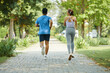 © DragonImages - Man and woman jogging together along a park path during a sunny day, both wearing athletic wear
