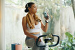 © DragonImages - A woman hydrating with a water bottle while sitting on an exercise bike after a workout session