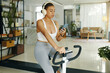 © DragonImages - A woman in workout clothes using her phone while exercising on a stationary bike in a bright living room