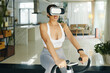 © DragonImages - A woman exercising on a stationary bike wearing a VR headset in a contemporary living room, exploring virtual reality fitness