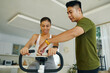© DragonImages - A man and woman adjusting the settings on an exercise bike together in a bright, modern living room focused on fitness