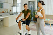 © DragonImages - A man and woman discussing their workout routine while the woman uses a stationary bike in a bright living space