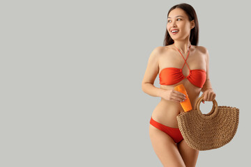 Sticker - Young Asian woman in swimsuit with sunscreen cream and bag on light background