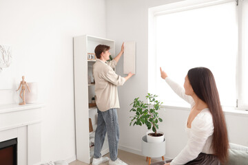 Poster - Young man hanging painting on light wall with his wife at home