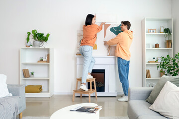 Young couple hanging paintings on light wall above fireplace at home, back view