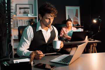 Wall Mural - Sitting man with serious face working on laptop waiting email for final project at night time while holding coffee cup to check overtime. Blurry secretary checking paper report background. Postulate.