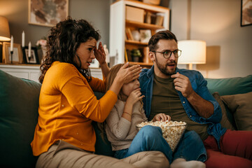 Wall Mural - Family of three holding a popcorn pot and watching a scary movie together in the living room