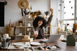 © sofiko14 - Young Caucasian woman potter happy with online sale of mug on Internet against background of shelves with ceramics. Curly-haired pretty business lady engaged in retail trade of handmade tableware.
