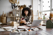 © sofiko14 - Owner of pottery workshop updates database of items that sold on website, sitting at table against background of shelves with products. Caucasian businesswoman engaged in filling social networks.