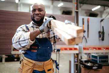 Male carpenter carrying wood planks on his shoulder. Multiracial woodworker wearing safety apron and gloves working in furniture builder and renovation in carpenter's shop. Joiner, repairman at work.