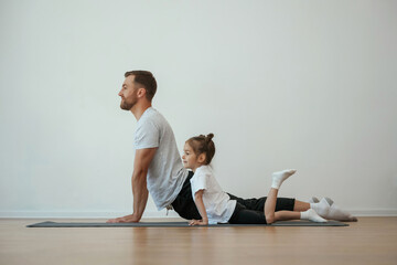 Wall Mural - Against white wall, stretching. Father with little daughter are doing yoga at home