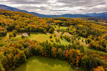 Wall Mural - Autumn forest adorns isolated farmhouse under clouds, mountain backdrop. Dron view. Vermont, USA.