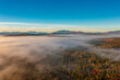 © romankrykh - Top drone view through clouds on multicolor trees in fog at dawn in autumn. Mountains background.