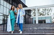 © Dragana Gordic - Two cheerful Caucasian women in medical attire with stethoscopes discussing joyfully as they leave a modern healthcare facility, embodying relief after a day's work.