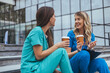 © Dragana Gordic - Two smiling Caucasian female nurses in scrubs relax with coffee on steps outside a medical facility, taking a well-deserved break from healthcare duties.