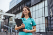 © Dragana Gordic - A joyful Caucasian female nurse stands confidently outside a hospital, holding a clipboard while dressed in teal scrubs with a stethoscope around her neck, showcasing professional healthcare